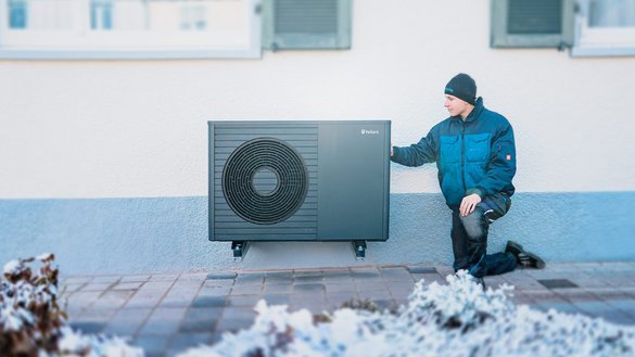Ein Techniker kniet neben einer modernen Wärmepumpe an einer Hauswand, um sie zu überprüfen oder zu installieren.