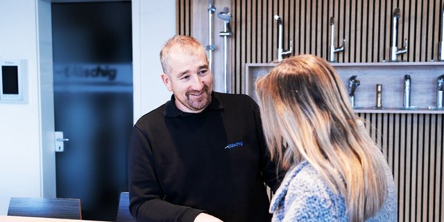 Ein Mann mit Bart lächelt freundlich, während er mit einer Frau in einem modernen Ausstellungsraum spricht.
