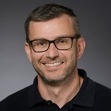 Ein lächelnder Mann mit Brille und kurzem Haar trägt ein schwarzes Poloshirt vor einem neutralen Hintergrund.