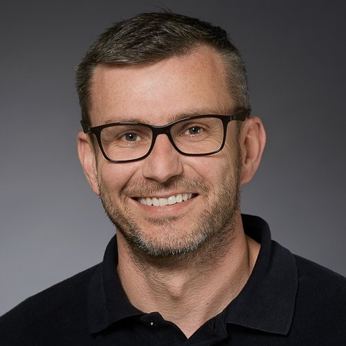 Ein lächelnder Mann mit Brille und kurzem Haar trägt ein schwarzes Poloshirt vor einem neutralen Hintergrund.